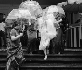 Bride in white dress descends steps in rain at The Columns Hotel, bridesmaids with clear umbrellas, dark dresses.
