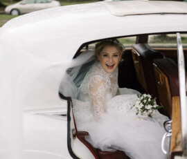 Bride in lace dress and veil smiles in a vintage white car with open door, bouquet in hand at The Columns Hotel wedding.