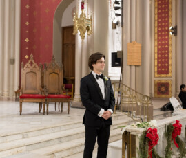 Smiling groom in black tuxedo stands at ornate church altar with red bows, greenery, and gold accents in New Orleans wedding.
