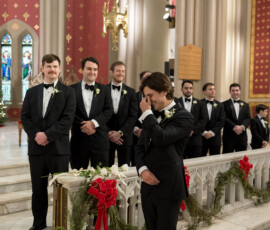 A groom wipes his eyes next to groomsmen in a decorated church with holiday greenery, red bows, and stained glass.