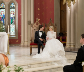 Bride and groom sit on ornate chairs in a formal New Orleans church wedding with red walls, stained glass, and gold accents.