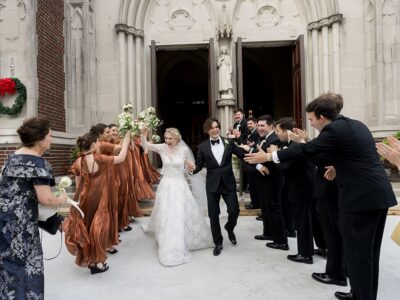 Bride and groom smile, walking hand in hand as bridal party cheers outside church at classic New Orleans wedding.