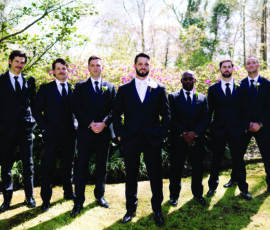 Groom and groomsmen dressed in black tuxedos posing outdoors in a garden setting, standing confidently in sunlight with blooming greenery behind them.