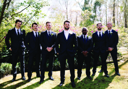 Groom and groomsmen dressed in black tuxedos posing outdoors in a garden setting, standing confidently in sunlight with blooming greenery behind them.