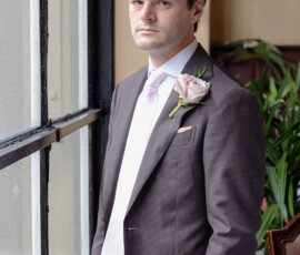 Man in dark suit with pink tie and rose boutonniere stands by window; plants and soft light evoke New Orleans wedding elegance.