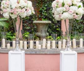 Glass vases of white and pale pink flowers on pedestals, with candles and stone fountain before a brick wall with greenery.