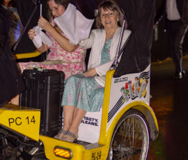 Two women in a yellow pedicab at night, one smiling, one covering her head, amid colorful lights after a Louisiana wedding.
