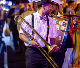 Man in white shirt, bow tie, and hat plays trombone in lively New Orleans-style nighttime parade, perfect for wedding inspiration.