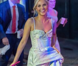 Smiling woman in a light blue dress walks at a night wedding, holding her skirt. Formal guests and colorful lights in background.