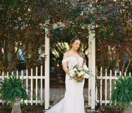 Bride in off-shoulder gown stands by white garden arch, holding flowers amid greenery at a sunny New Orleans wedding.
