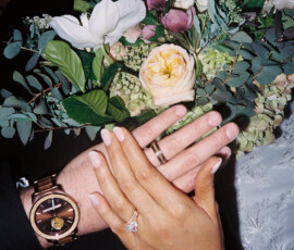 Hands with wedding rings rest on a white, pink, and yellow bouquet at a Louisiana wedding; manicure and watch visible.
