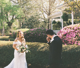 Bride in white dress smiles holding bouquet as groom wipes eyes by gazebo and blooming bushes at Louisiana wedding.