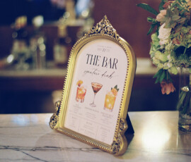 Gold-framed bar menu with cocktail illustrations on marble next to white and peach flowers—elegant New Orleans wedding inspo.