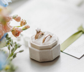 Hexagonal velvet ring box with diamond and gold rings on white surface, pastel flowers, and green ribbon for Gulf South wedding inspo.