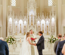 Bride and groom hold hands at a Louisiana church altar as a priest officiates. Flowers, candles, and guests are present.