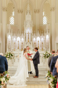 Bride and groom hold hands at a Louisiana church altar as a priest officiates. Flowers, candles, and guests are present.