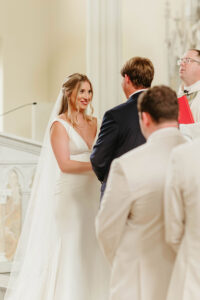 Bride in white dress smiles at groom during Gulf South church wedding, with officiant and guests in foreground.