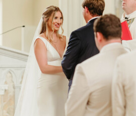 Bride in white dress smiles at groom during Gulf South church wedding, with officiant and guests in foreground.