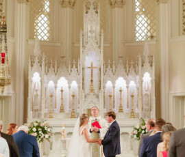 Bride and groom hold hands at a grand Louisiana-inspired church wedding, with guests and priest before an ornate white altar backdrop.