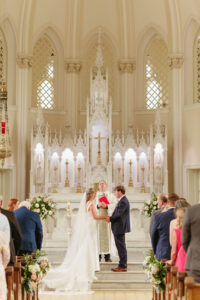 Bride and groom hold hands at a grand Louisiana-inspired church wedding, with guests and priest before an ornate white altar backdrop.