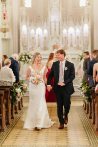 Smiling bride and groom walk hand in hand down the church aisle after their Louisiana wedding, surrounded by guests.