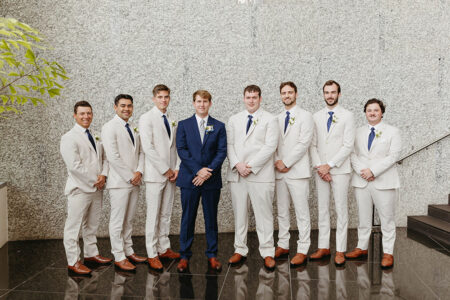 Eight men indoors—one in a navy suit centers seven in cream suits with blue ties on a black floor by a light stone wall.