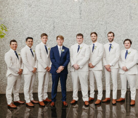 Eight men indoors—one in a navy suit centers seven in cream suits with blue ties on a black floor by a light stone wall.