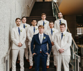 Groom in navy suit with seven groomsmen in cream suits, blue ties, indoors at elegant New Orleans wedding with granite walls.