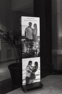 Digital screen with three black-and-white engagement photos and “Congratulations: Rory & Brennan - 8.9.23” in indoor venue.