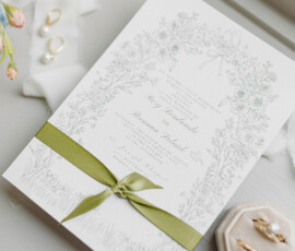 Floral wedding invitation with green ribbon, gold rings, pearl earrings, and blue flowers on a table—New Orleans elegance.