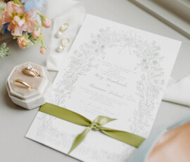 Elegant wedding invitation with floral designs, green ribbon, rings, pastel flowers, and jeweled blue shoe on table.
