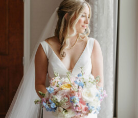 Bride in white dress with pastel bouquet stands by window, light streaming in, awaiting her New Orleans wedding ceremony.