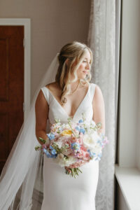 Bride in white dress with pastel bouquet stands by window, light streaming in, awaiting her New Orleans wedding ceremony.
