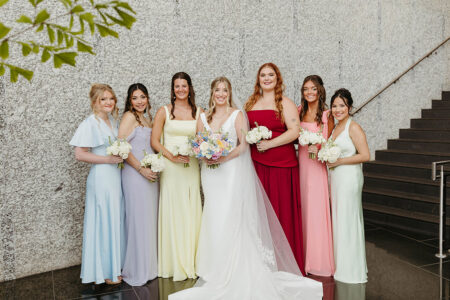 Bride and six bridesmaids in colorful dresses pose indoors at a Louisiana wedding, with stairs and greenery behind them.