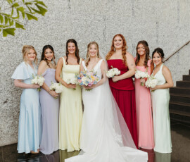 Bride and six bridesmaids in colorful dresses pose indoors at a Louisiana wedding, with stairs and greenery behind them.
