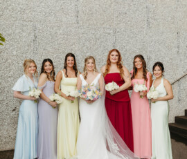 Bride in a white gown and six bridesmaids in pastel dresses pose with bouquets by a stone wall at their Louisiana wedding.