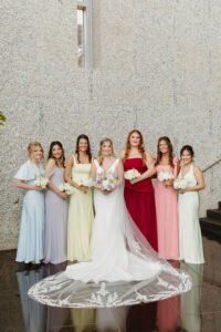 Bride in a white gown and six bridesmaids in pastel dresses pose with bouquets by a stone wall at their Louisiana wedding.