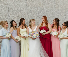 Seven women in colorful dresses pose with bouquets by a stone wall at a New Orleans wedding; the bride wears white.