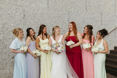 Seven women in colorful dresses pose with bouquets by a stone wall at a New Orleans wedding; the bride wears white.