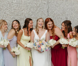 Bride in white with six bridesmaids in pastel and red dresses, laughing by a stone wall with bouquets—Gulf South wedding inspiration.
