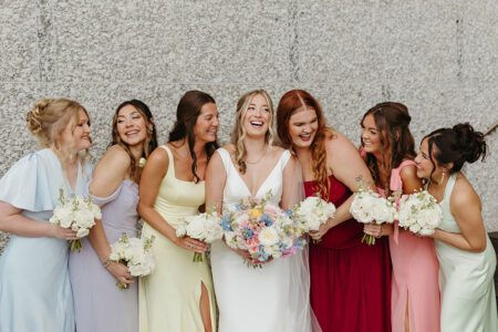 Bride in white with six bridesmaids in pastel and red dresses, laughing by a stone wall with bouquets—Gulf South wedding inspiration.