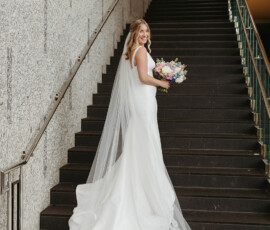 Bride in white gown and long embroidered veil stands on staircase, smiling with bouquet before her Gulf South wedding.