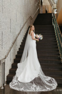 Bride in white gown and long embroidered veil stands on staircase, smiling with bouquet before her Gulf South wedding.