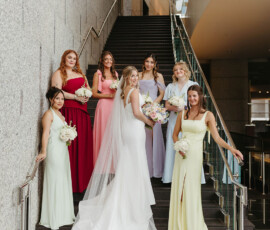 Bride in white gown with six bridesmaids in pastel and red dresses on a modern staircase at a New Orleans wedding, smiling.