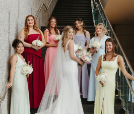 Bride in white with six bridesmaids in pastel and red dresses on stairs, all smiling and holding bouquets at New Orleans wedding.