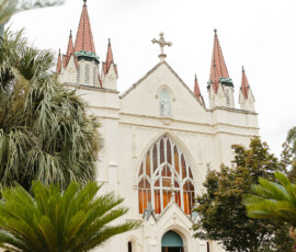 White Gothic church with red spires, arched stained glass, palm trees, and flowers—romantic Louisiana wedding backdrop.