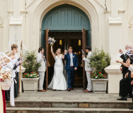 Smiling bride and groom exit a New Orleans church as guests wave ribbons on flower-lined steps.
