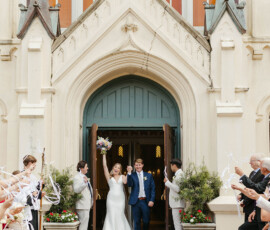 Newlyweds leave a church smiling, holding hands as guests cheer and wave streamers, radiating joy and elegance.