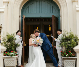 Bride and groom kiss on church steps, smiling friends with streamers nearby, surrounded by flowers at a New Orleans wedding.