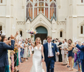 Newlyweds walk outside a church as guests wave streamers. Bride holds bouquet, groom smiles—classic New Orleans wedding.